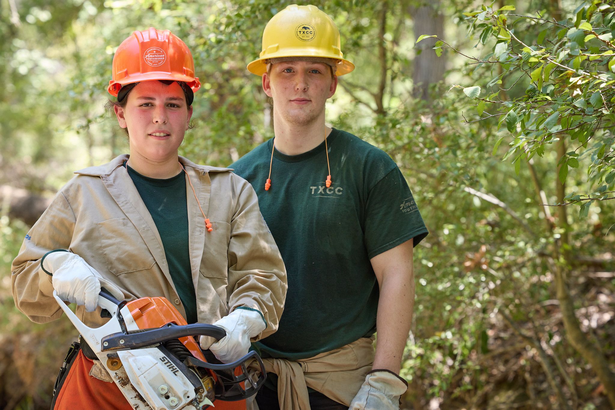 Two Americorps members pose in safety gear in a wooded outdoor environment.