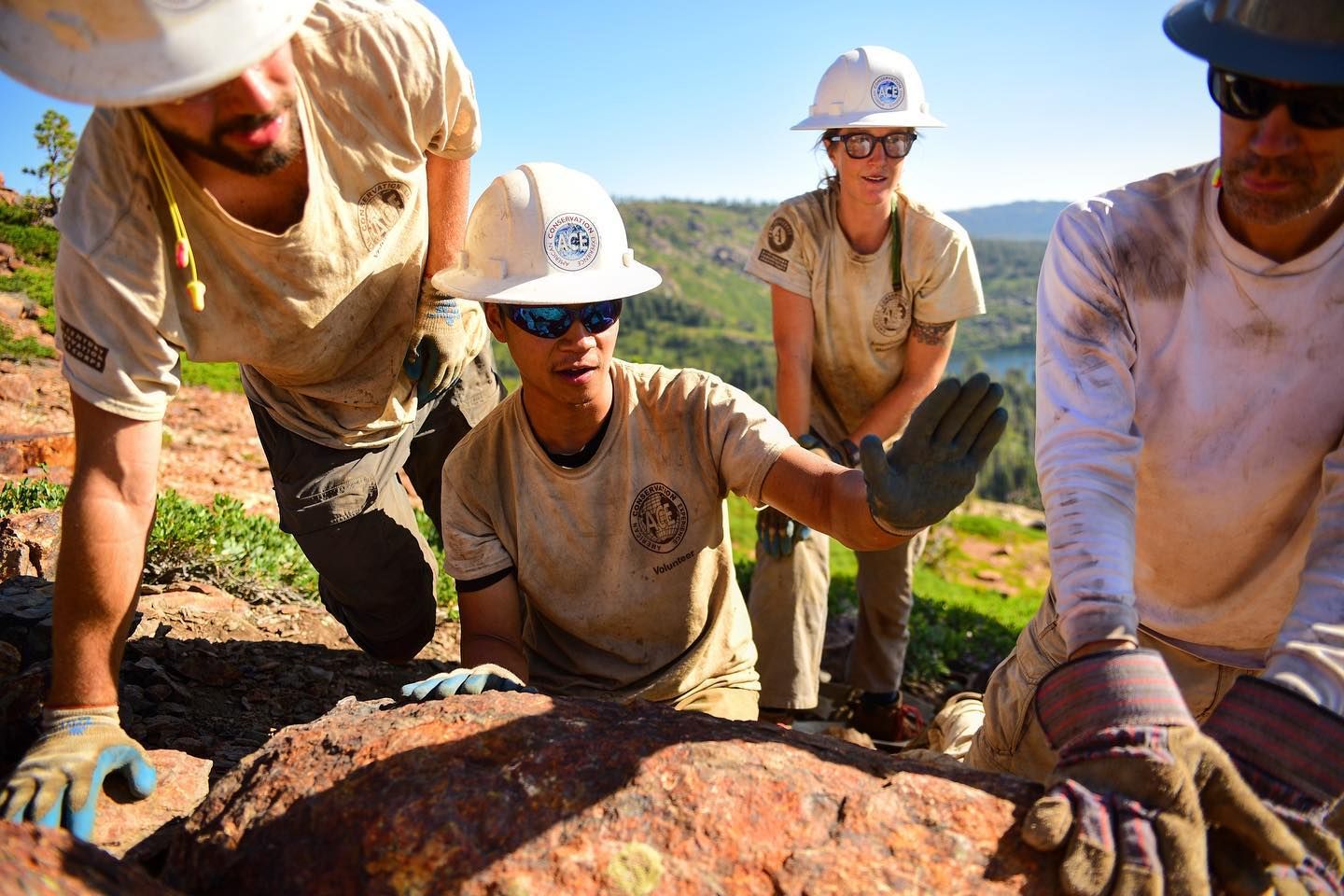 Four Americorps members in hard hats, dirty t-shirts, and work gloves work together outdoors in a rocky landscape.