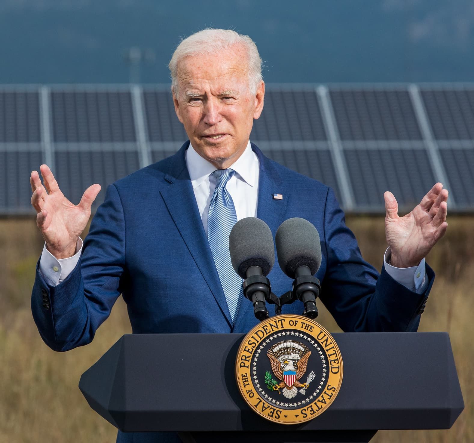 President Joe Biden speaking from behind a podium outdoors. Behind him is an array of solar panels.